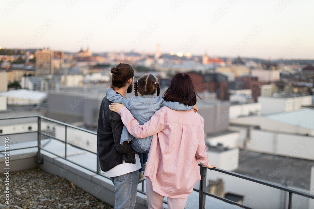 Fototapeta premium Caucasian family with young daughter standing on rooftop enjoying beautiful cityscape at sunset. Rear view of father, mother, and child embraced together.