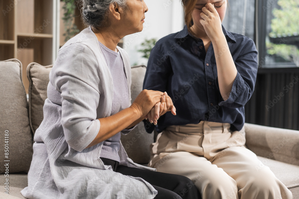 A mature mom and her young daughter, both Asian, sit on a sofa. The daughter appears sick and unwell, causing worry and possibly depression. The mom tries to encourage her and stay by her side.