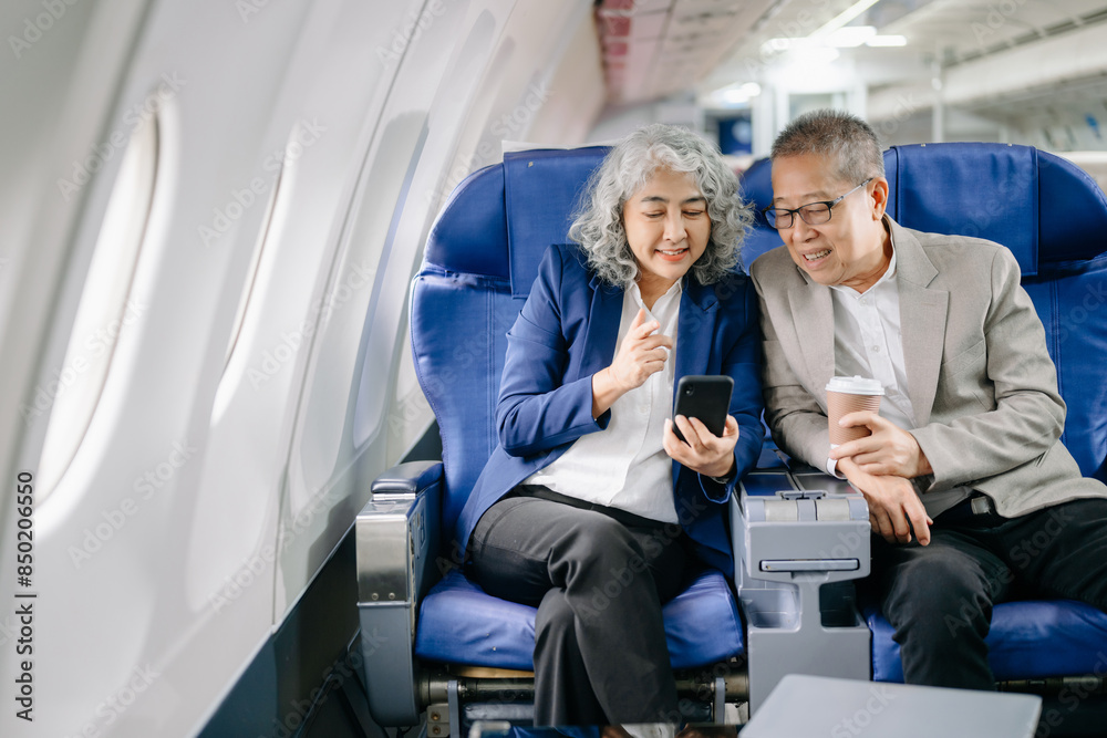 Smiling senior couple of Asian tourists take photos of each other while sitting on a plane. with digital tablet, laptop and smartphone. Travel in style, work Smiling senior couple of Asian tourists take photos of each other while sitting on a plane. with digital tablet, laptop and smartphone. Travel in style, work