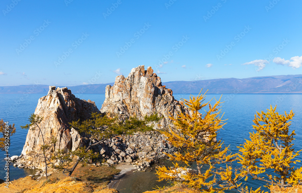 Obraz premium Lake Baikal in autumn. The famous Shamanka rock framed by yellowed larches on a sunny October morning. Traveling around the autumn island of Olkhon. Picturesque landscape. Natural background
