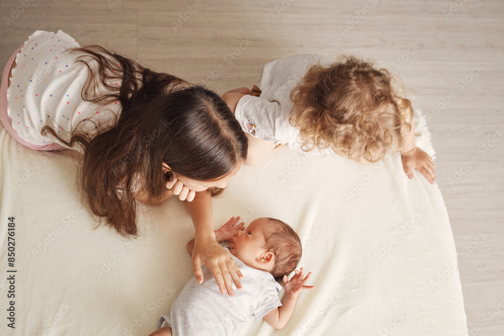 Top view of sisters in their casual outfits gently stroking their baby brother's tiny hands and feet enjoying him sitting in a warm bedroom meeting with new family member Top view of sisters in their casual outfits gently stroking their baby brother's tiny hands and feet enjoying him sitting in a warm bedroom meeting with new family member