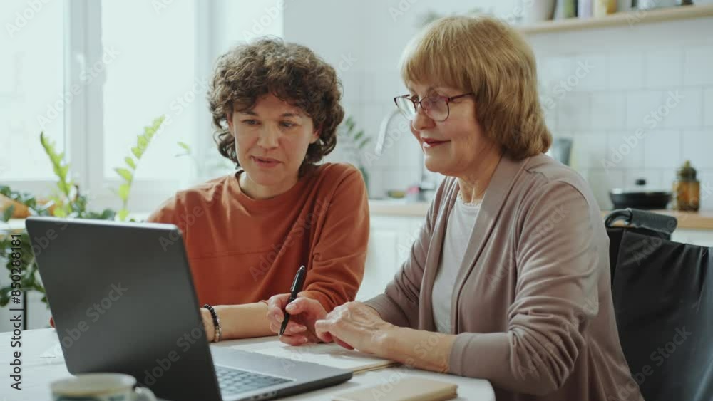 Young woman using laptop and explaining how to use online banking to senior grandmother sitting in wheelchair and taking notes during the day at home Young woman using laptop and explaining how to use online banking to senior grandmother sitting in wheelchair and taking notes during the day at home
