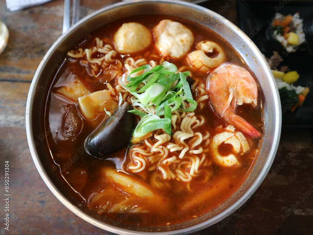 a bowl of noodles or ramyeon with a mixture of green mussels, shrimp, fish balls, meatballs with red tom yum-like. sauce. Korean noodles. Ramen street food. a bowl of noodles or ramyeon with a mixture of green mussels, shrimp, fish balls, meatballs with red tom yum-like. sauce. Korean noodles. Ramen street food.