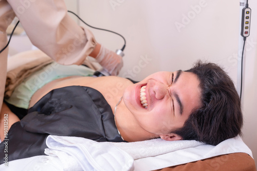 A man grimaces in pain while undergoing a Radio Frequency body sculpting treatment at an aesthetic clinic.