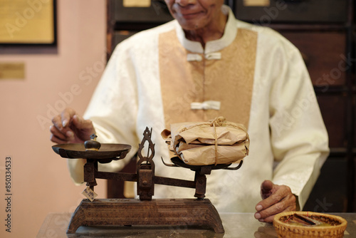 Obraz na plátně Senior apothecary in traditional Chinese clothes measuring packed herbs on scale