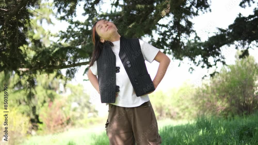 Asian girl is doing exercise outdoors, standing under a large tree. She wears a white t-shirt and a black vest, performing stretches on the green grass Asian girl is doing exercise outdoors, standing under a large tree. She wears a white t-shirt and a black vest, performing stretches on the green grass