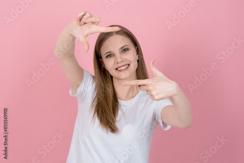 Attractive caucasian young blonde woman in casual white t-shirt looking at camera, smiling and showing hands gesture camera frame isolated on pink studio background.