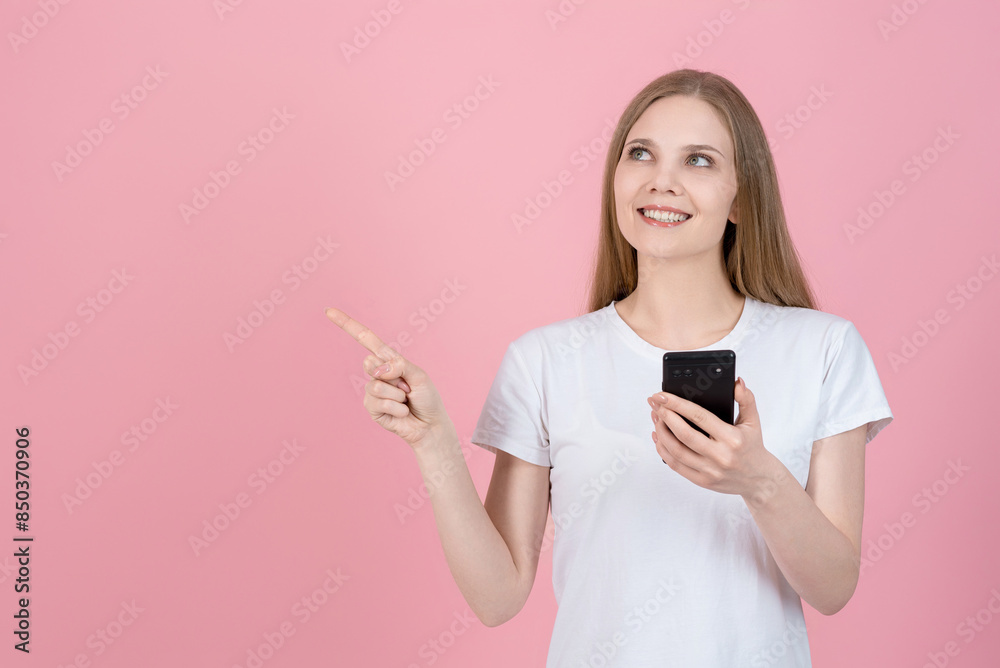 Attractive caucasian young blonde woman in casual white t-shirt using mobile phone and pointing finger at empty copy space for advertising isolated on pink studio background.