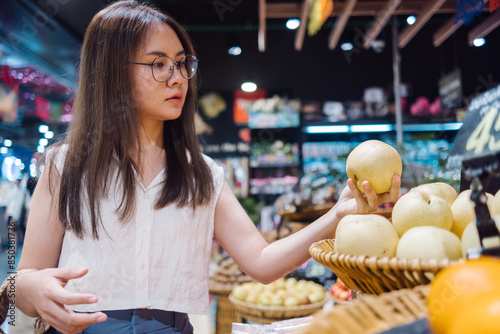 Woman in a grocery store holding a pear, surrounded by baskets of fresh fruit. Suitable for designs focusing on everyday shopping, healthy eating, or retail promotions.