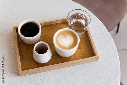 Coffee set on white cafe table. Cappuccino with heart shaped latte art, black filer drip coffee, espresso and water in cafeteria. Alternative brewing, morning breakfast. Minimal concept background
