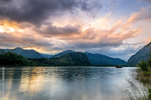 Fotografie A picturesque scene at Almsee Lake in Austria, featuring a calm lake reflecting a golden sunset, with a small cabin nestled in the water and towering mountains in the background