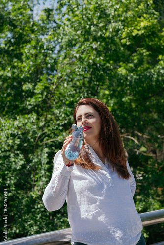Wallpaper Mural Mature 45 year old woman with long hair and red lips drinking water from a bottle in nature on a sunny day. Copy space.  Healthy lifestyle concept Torontodigital.ca