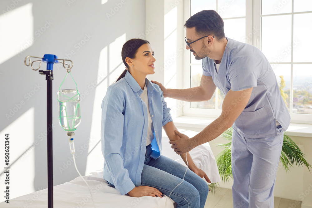 Young male nurse or man doctor giving consultation to his female smiling patient sitting on the couch in medical clinic and receiving IV drip infusion and vitamin injection therapy. Young male nurse or man doctor giving consultation to his female smiling patient sitting on the couch in medical clinic and receiving IV drip infusion and vitamin injection therapy.