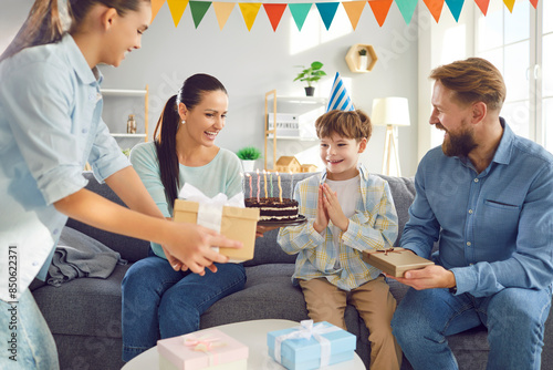 Happy family sitting on sofa in living room at home and congratulating their smiling cute child boy on birthday party giving him cake and present gift boxes. Holiday celebrating concept.