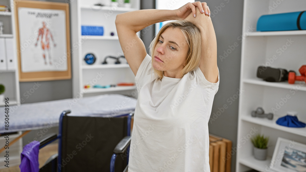 A young blonde woman stretching her arm in a bright rehabilitation clinic room. A young blonde woman stretching her arm in a bright rehabilitation clinic room.
