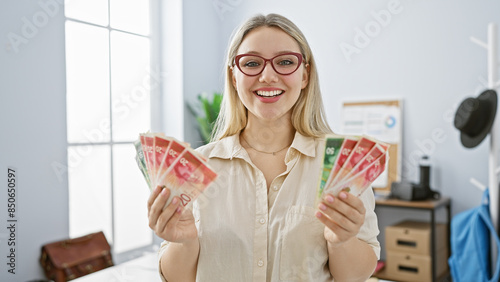 Schilderij op canvas Blonde woman in glasses smiling while holding israeli shekels in a bright office interior