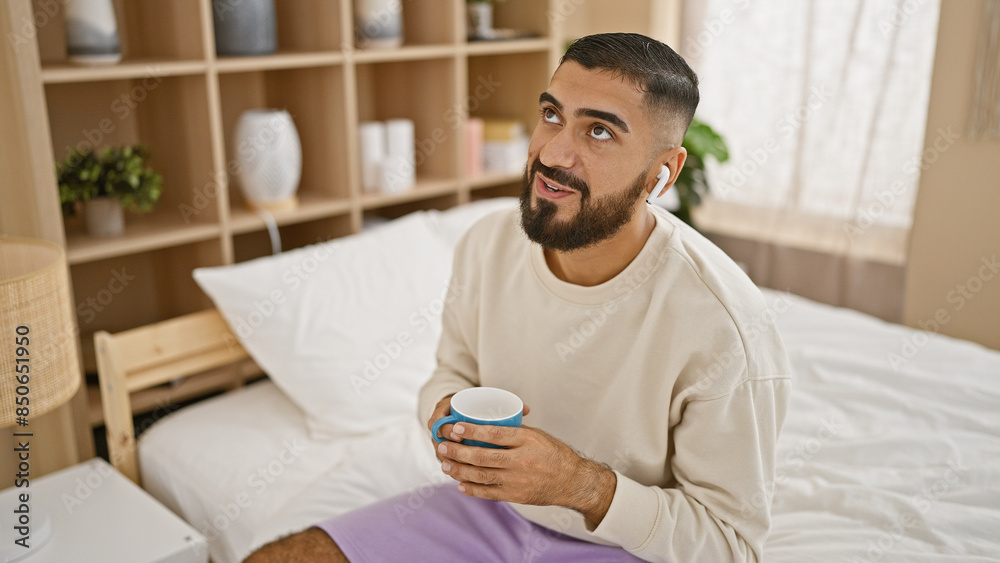 A relaxed young man with a beard sitting in a bedroom, enjoying a coffee and wearing earphones. A relaxed young man with a beard sitting in a bedroom, enjoying a coffee and wearing earphones.