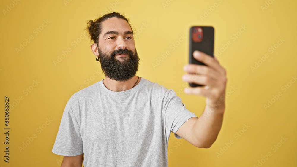 Handsome hispanic man with a beard taking a selfie against a yellow wall.