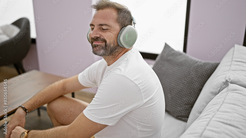 Fototapeta premium Relaxed middle-aged man with beard smiling, wearing headphones and white shirt, sitting on a couch indoors.