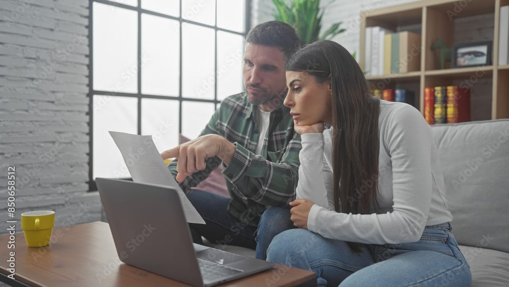 A hispanic man and woman reviewing documents together on a sofa indoors with a laptop and cup nearby. A hispanic man and woman reviewing documents together on a sofa indoors with a laptop and cup nearby.