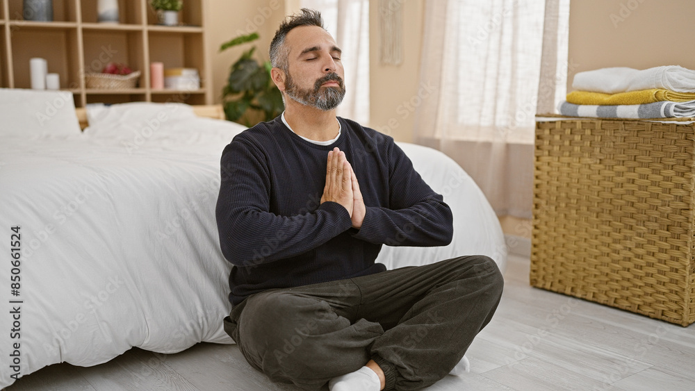 Fototapeta premium Mature hispanic man meditates with closed eyes, beard grey, in bedroom interior, portraying tranquility and wellness.