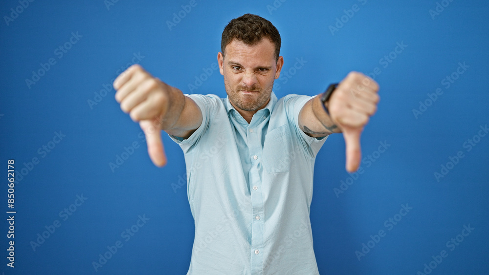 Portrait of a young man with a beard giving two thumbs down against a blue wall outdoors. Portrait of a young man with a beard giving two thumbs down against a blue wall outdoors.