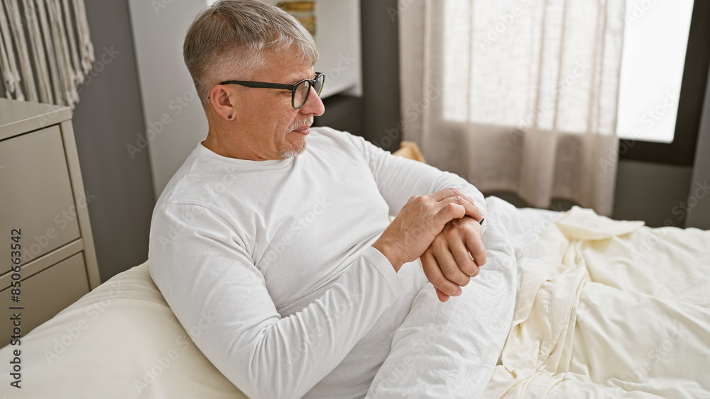 Mature grey-haired man in white shirt relaxing on bed in a well-lit bedroom interior Mature grey-haired man in white shirt relaxing on bed in a well-lit bedroom interior