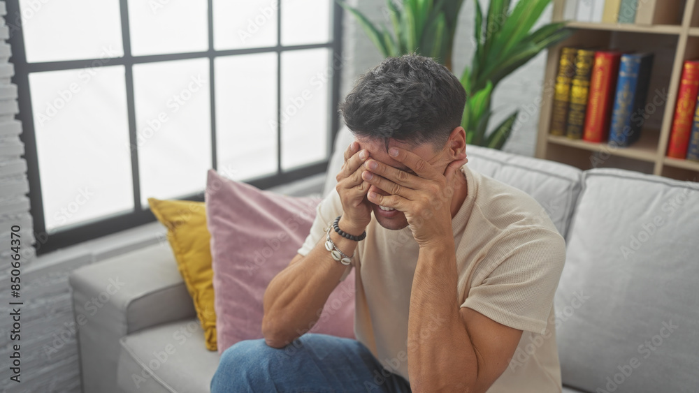 A distressed hispanic man covering his face with hands while sitting on a sofa indoors. A distressed hispanic man covering his face with hands while sitting on a sofa indoors.