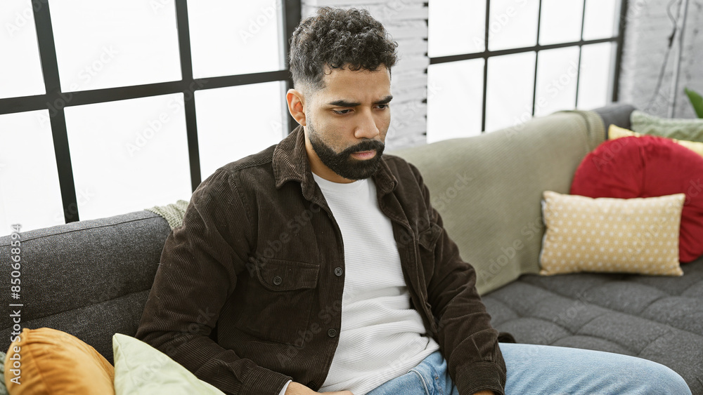 Thoughtful young man with beard sitting on couch at home looking pensive Thoughtful young man with beard sitting on couch at home looking pensive
