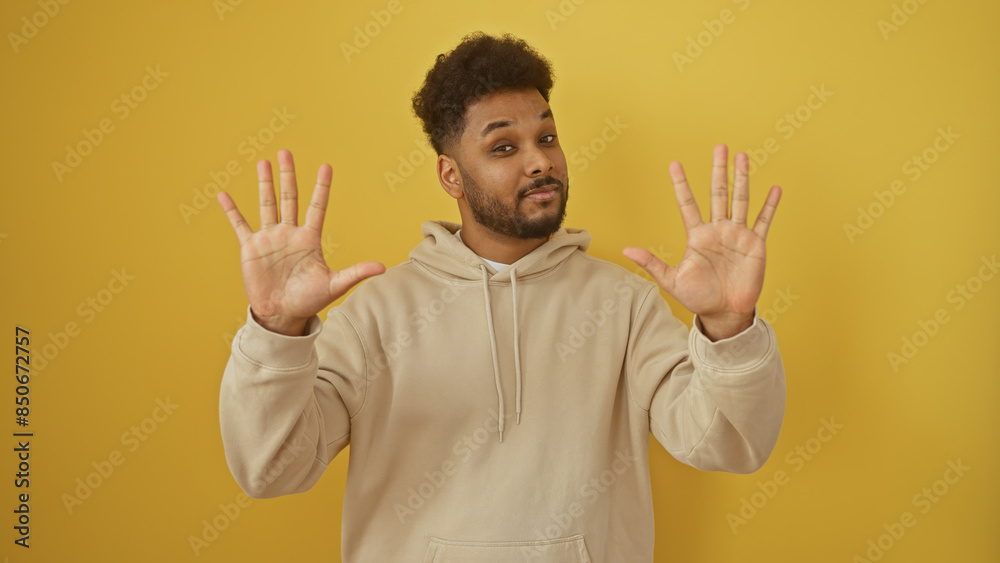 Handsome man showing palms in a casual hoodie against a vibrant yellow background, expressing openness.