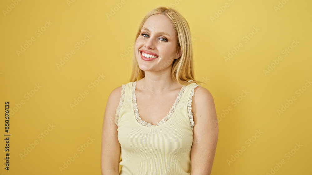 Obraz premium A smiling young caucasian woman with blonde hair wearing a sleeveless top poses against an isolated yellow background.