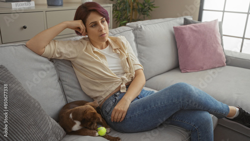 A young caucasian woman lounges thoughtfully on a grey sofa in her apartment living room, with a small dog resting near her, highlighting a serene indoor setting with casual home decor. © Krakenimages.com