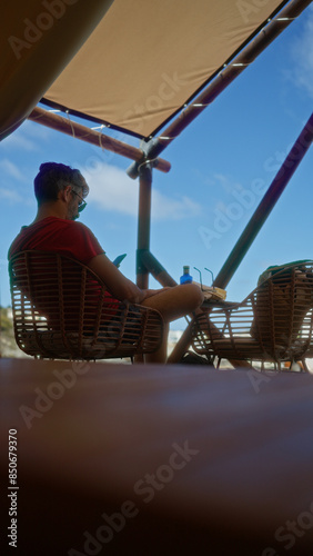 Man relaxing under a tent in african nature with a phone in hand, surrounded by adventure and camping gear.