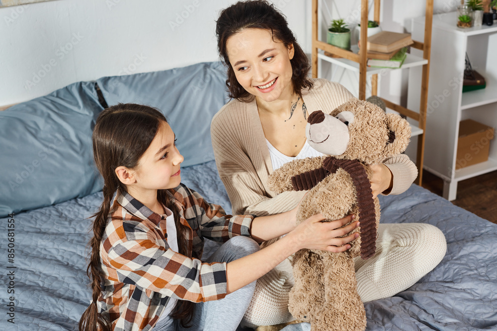 A mother tenderly holds a teddy bear while sitting next to her daughter on a cozy bed. A mother tenderly holds a teddy bear while sitting next to her daughter on a cozy bed.