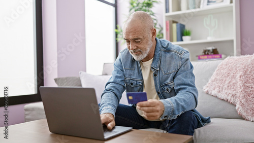 Senior man with beard using laptop and holding credit card in a modern living room. © Krakenimages.com