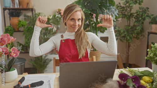 A young blonde woman in a red apron points to herself in a flower shop surrounded by plants and a laptop. © Krakenimages.com