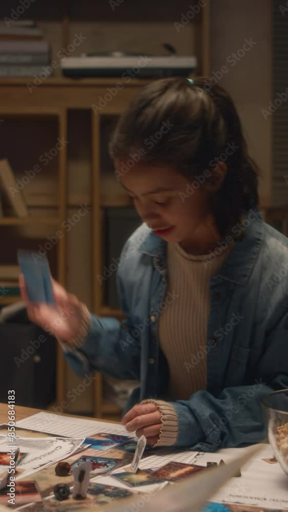 Vertical shot of Hispanic girl in denim jacket choosing card and rolling dice while playing table game with friends at home in evening Vertical shot of Hispanic girl in denim jacket choosing card and rolling dice while playing table game with friends at home in evening