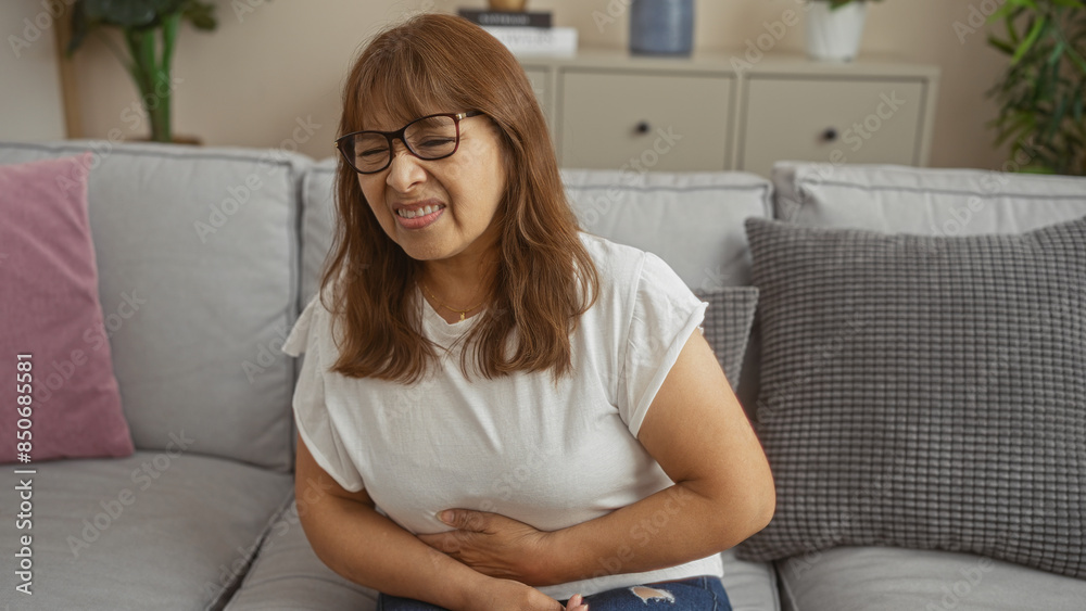 Middle-aged hispanic woman in pain sitting on a couch in a living room with a pained expression, clutching her stomach with decor visible. Middle-aged hispanic woman in pain sitting on a couch in a living room with a pained expression, clutching her stomach with decor visible.