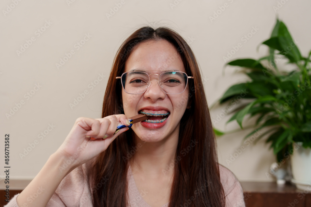 Asian girl with glasses enjoying a snack, smiling. Close-up of her biting a chocolate bar. Casual home setting with neutral background. Emphasis on cheerful expression and enjoying a sweet treat. Asian girl with glasses enjoying a snack, smiling. Close-up of her biting a chocolate bar. Casual home setting with neutral background. Emphasis on cheerful expression and enjoying a sweet treat.