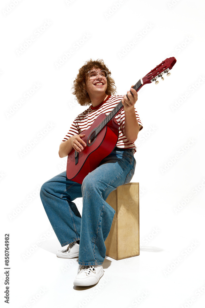Portrait of happy young man, musician wearing casual clothes, sitting on wooden box and playing guitar isolated on white background. Concept of music, festival, concert, hobby, talent, performance Portrait of happy young man, musician wearing casual clothes, sitting on wooden box and playing guitar isolated on white background. Concept of music, festival, concert, hobby, talent, performance