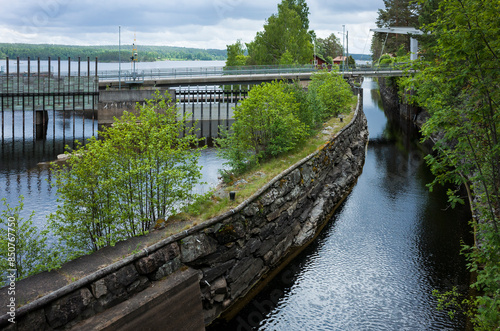 Narrow water channel with a stone wall separating it from the lake with a dam and bridge, green banks, cloudy weather, Dalsland Canal in Lennartsfors, Sweden