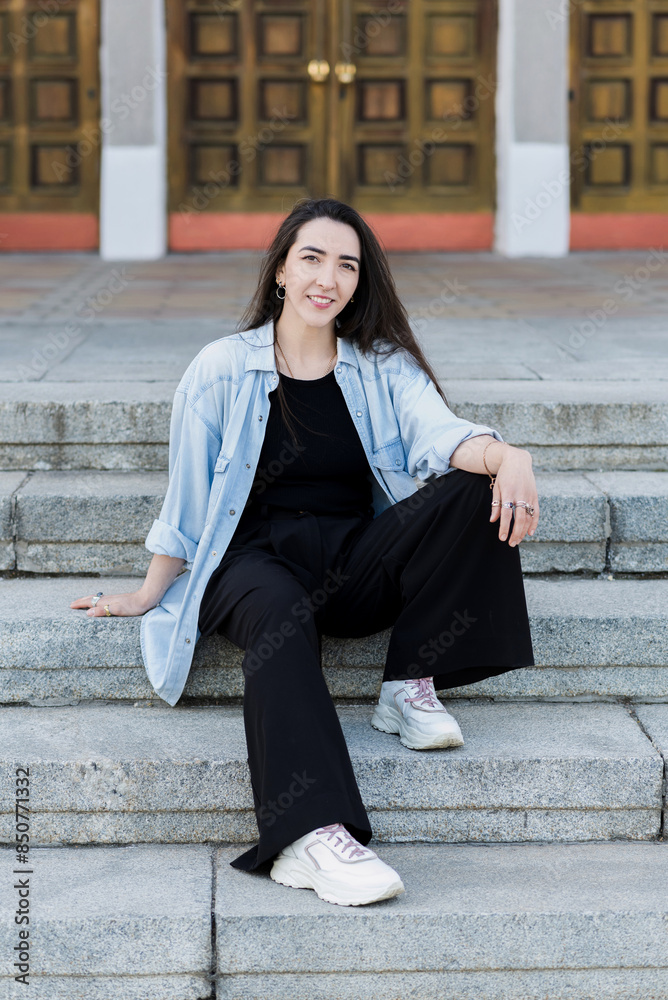 Attractive Caucasian Young Woman Brunette Hippie In Casual Clothes Posing With Pleasure While Walking Outdoors On A Warm Spring Day Sitting On The Steps. Stylish girl.