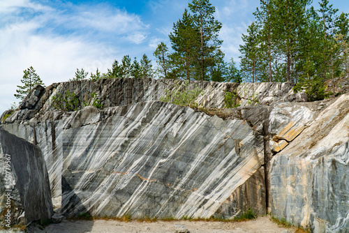 Scenic view of a marble quarry with distinct striations and pine trees in the background. The image highlights the contrast between the natural stone formations and the greenery under a bright blue