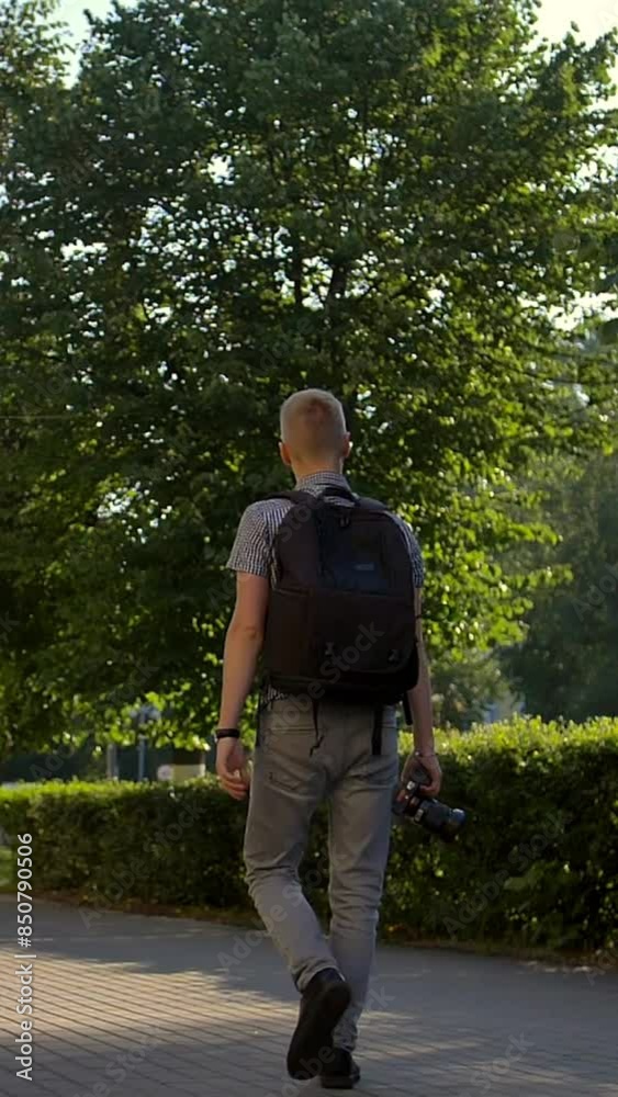 Young photographer is walking along a city street behind him. He looks around, looking for interesting sights. Towards natural sunlight Dawn. Wide shot in slow motion. Vertical shot. Young photographer is walking along a city street behind him. He looks around, looking for interesting sights. Towards natural sunlight Dawn. Wide shot in slow motion. Vertical shot.