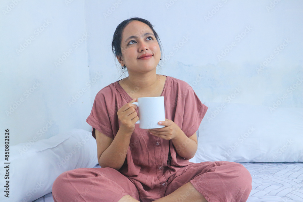 Smiling asian woman in pink pajamas in bed holding cup at morning. Enjoying weekend