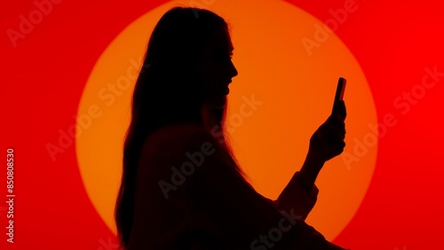 Stylish young woman black silhouette in casual talking by video call on smartphone online. Isolated on red studio background spotlight. photo