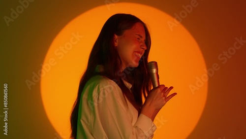 Stylish young woman in casual clothing singing son in karaoke microphone. Isolated on colorful studio background yellow spotlight. photo