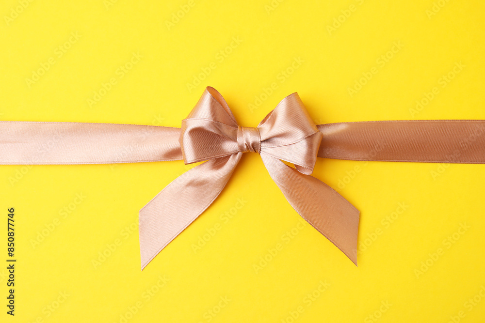 Fototapeta premium Beige satin ribbon with bow on yellow background, top view