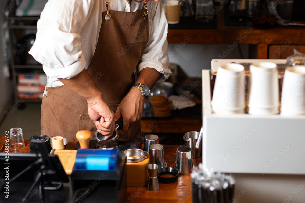 A barista in a brown apron tamping espresso grounds in a busy café setting, highlighting the meticulous coffee-making process and the art of crafting quality beverages. A barista in a brown apron tamping espresso grounds in a busy café setting, highlighting the meticulous coffee-making process and the art of crafting quality beverages.