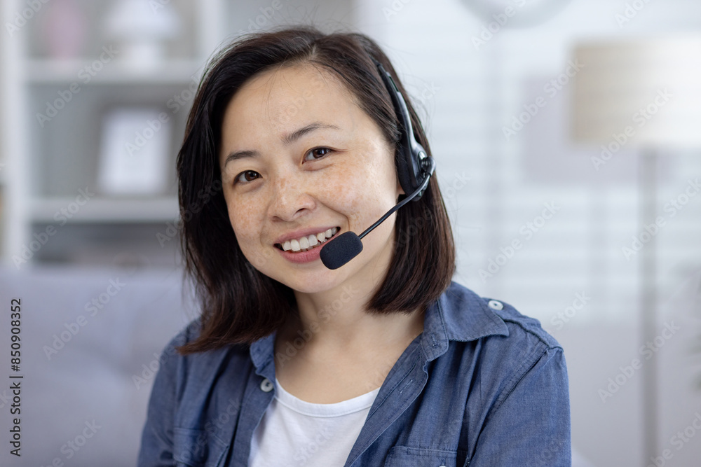 Close-up portrait of a young Asian woman sitting in front of the camera in her home office wearing a headset and smiling. Close-up portrait of a young Asian woman sitting in front of the camera in her home office wearing a headset and smiling.